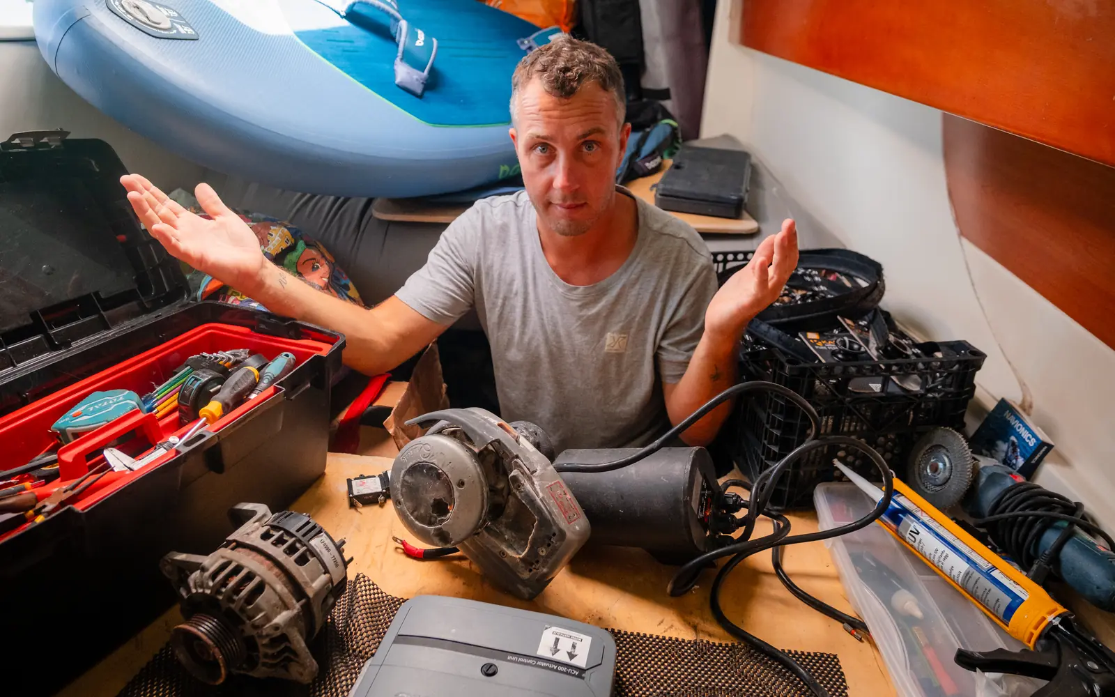 A sailor in the cockpit locker of a sailing boat shrugs among an alternator, an autopilot control unit, an open toolbox and a tube of sealant.